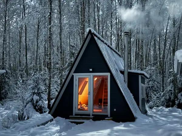 A small, dark A-frame cabin with a smoking chimney glows warmly from within, nestled in a dense, snow-covered forest.