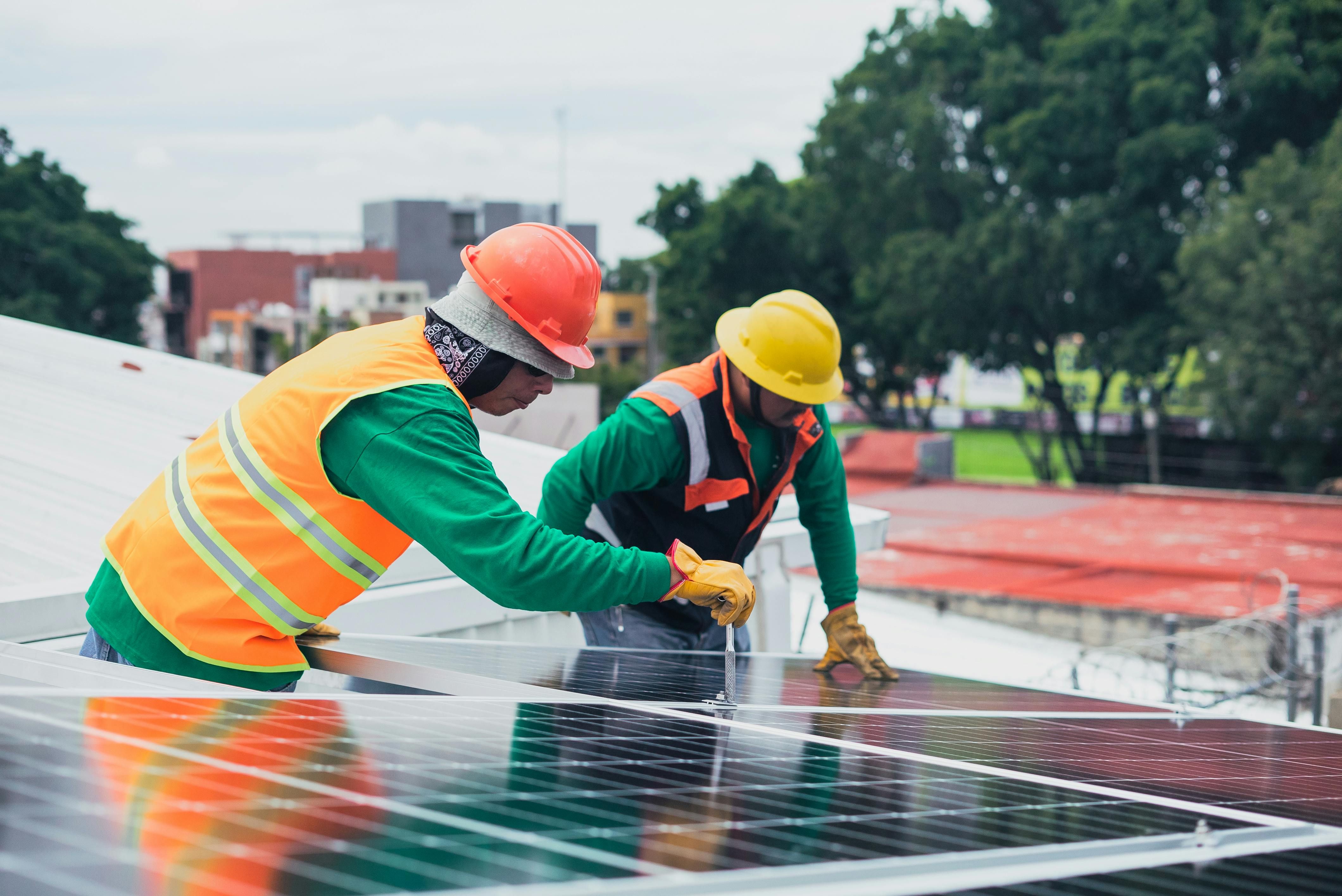 Two construction workers wearing hard hats and high-visibility vests install solar panels on a rooftop.