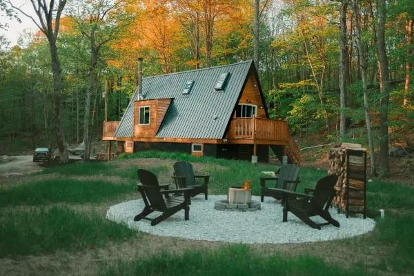 A wooden A-frame house with a metal roof and side dormer overlooks a gravel fire pit area with black Adirondack chairs set against a backdrop of autumn trees.