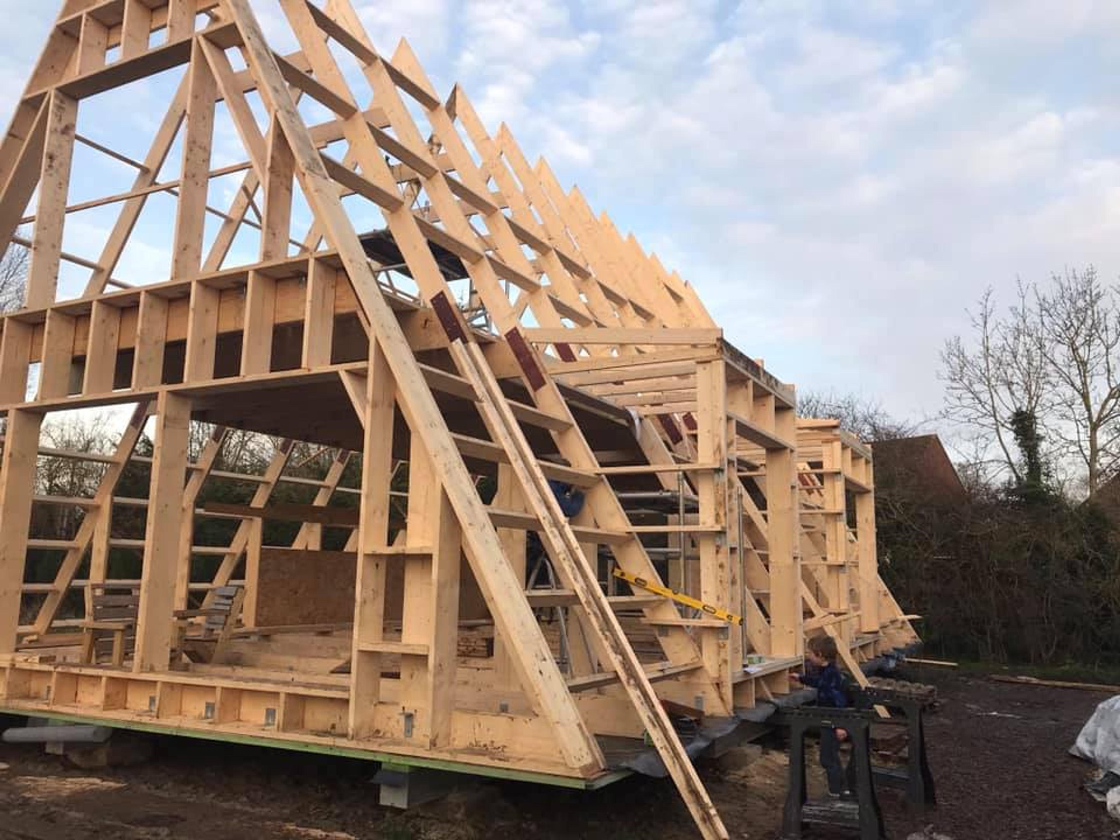 The complex wooden structural framing of a large A-frame house is shown under construction with visible rafters and wall studs against a cloudy sky.
