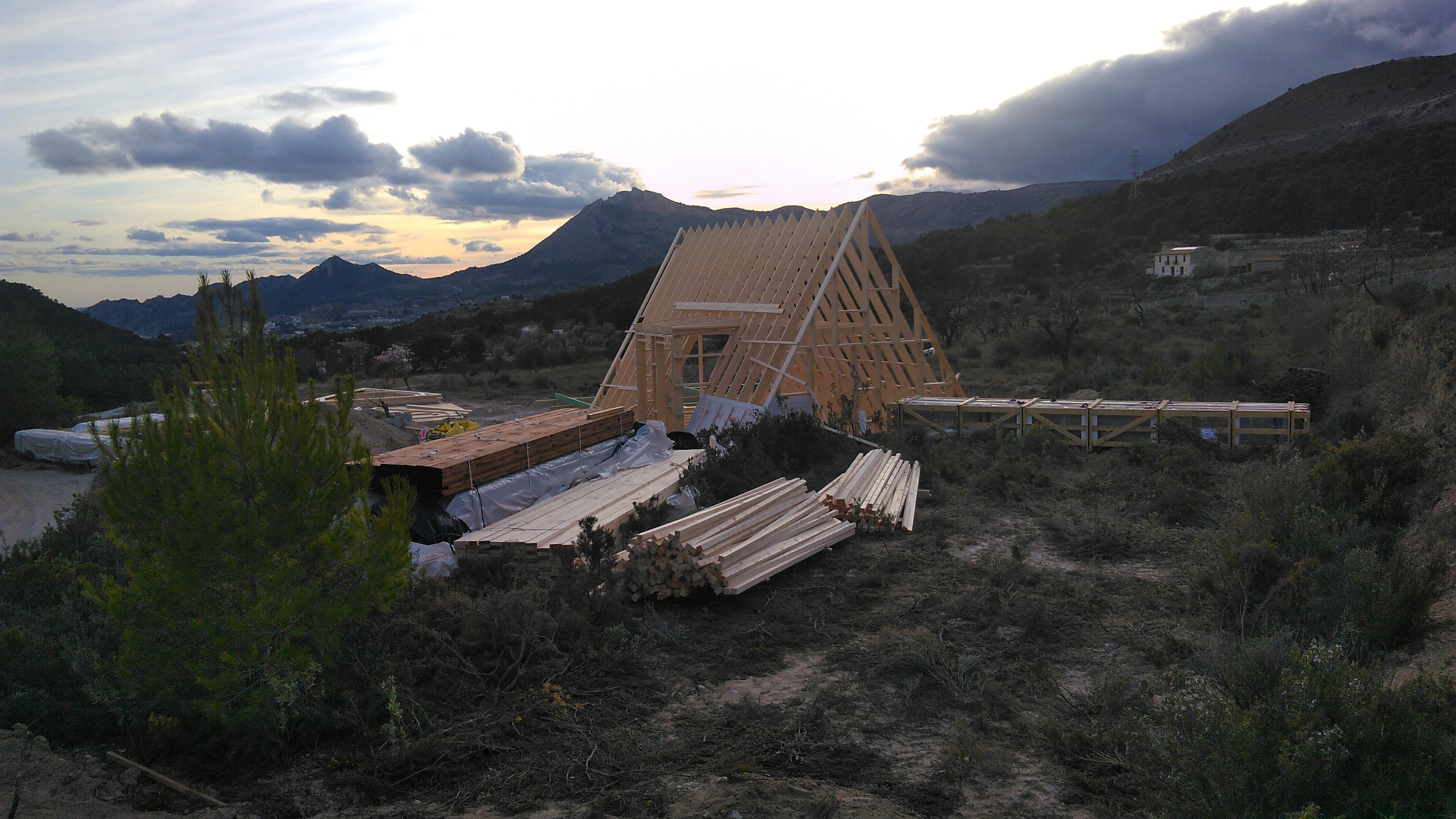 The structural wooden framework of an A-frame house kit under construction on an open hillside lot. Stacks of pre-cut timber and lumber sit organized in the foreground, with rolling mountain ranges and a cloudy sunset visible in the distance.