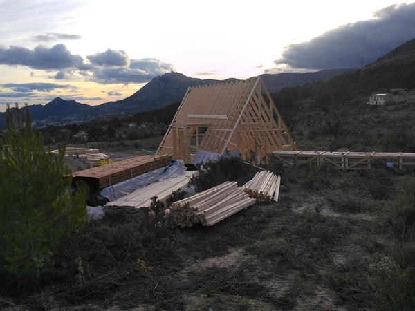 The structural wooden framework of an A-frame house kit under construction on an open hillside lot. Stacks of pre-cut timber and lumber sit organized in the foreground, with rolling mountain ranges and a cloudy sunset visible in the distance.