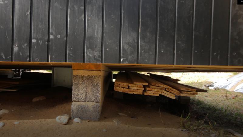A low-angle view shows the concrete block foundation and floor joists of a black-sided Trio 57 model, with extra lumber stored in the crawl space.
