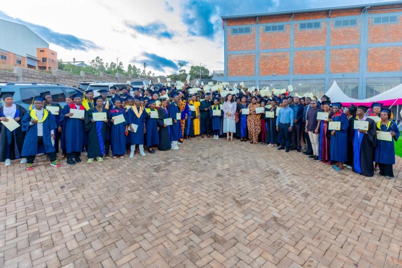 Dozens of DIKAM Training Center graduates in caps and gowns holding certificates in front of the factory