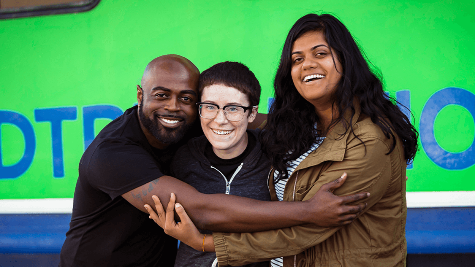 Three young adults embrace in front of Roadtrip Nation's green RV.