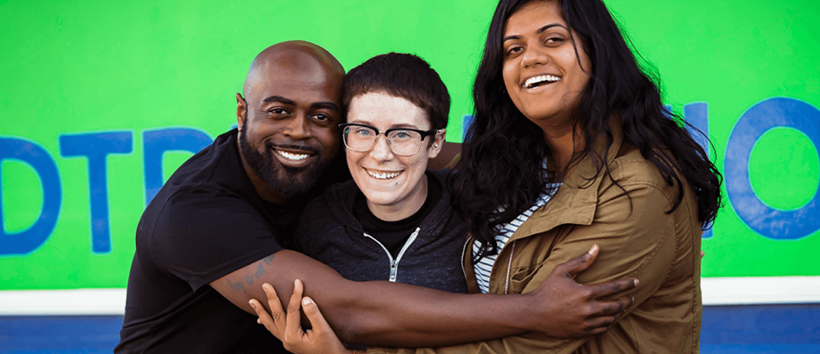 Three young adults embrace in front of Roadtrip Nation's green RV.