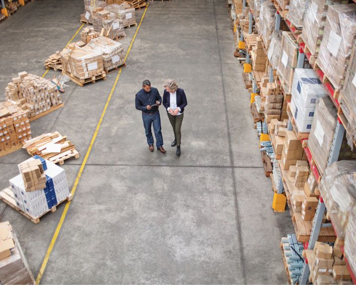 Two supply chain workers walking through a warehouse surrounded by packaged product. 