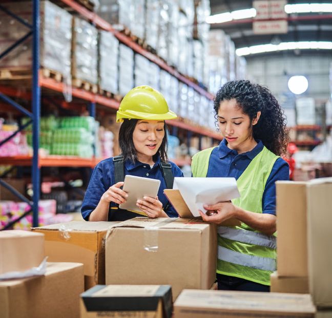 Warehouse workers go over inventory list. Boxes in foreground, warehouse in background. Women in supply chain.