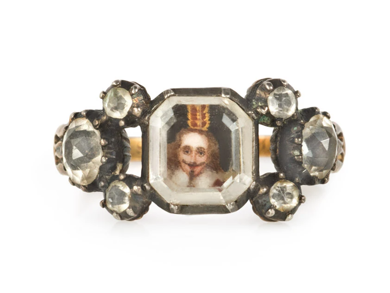 An antique Stuart crystal ring centering on a flat octagonal quartz revealing a miniature royal portrait, flanked on both sides by three old cut quartz and gold foliate shoulders, with engraved upper shank and under gallery, in 18k. Inscribed W. Mc 1738