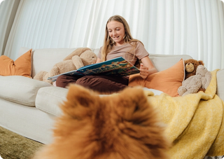 Pomeranian looking at girl on sofa reaching a book