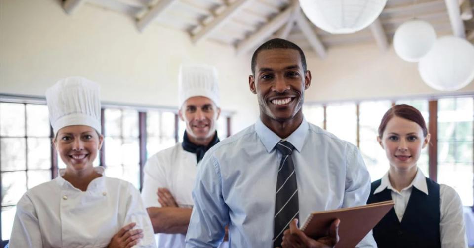 Four hospitality workers (two chefs, a manager holding a folder and a server) smile in a bright dining room.