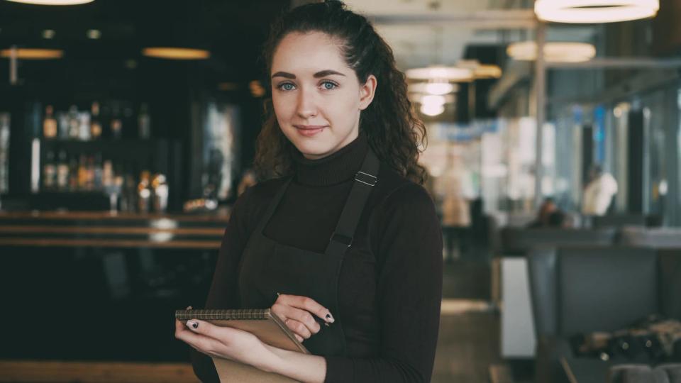 A smiling server in a dark apron holds a notepad in a restaurant, with a bar behind her.
