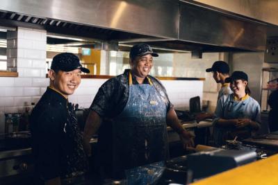 A quick-service kitchen crew in caps and aprons laughing together at the pass