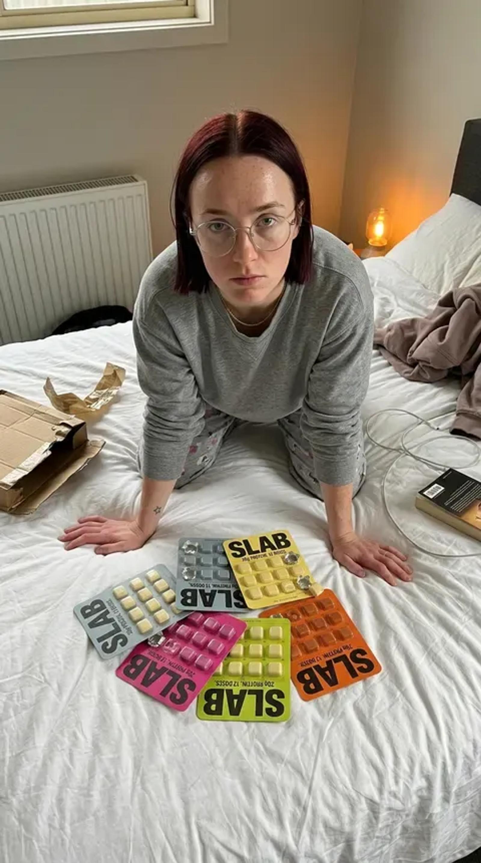 A woman kneeling on an unmade bed surrounded by SLAB trays in six different colourways, spelling the name across the duvet.