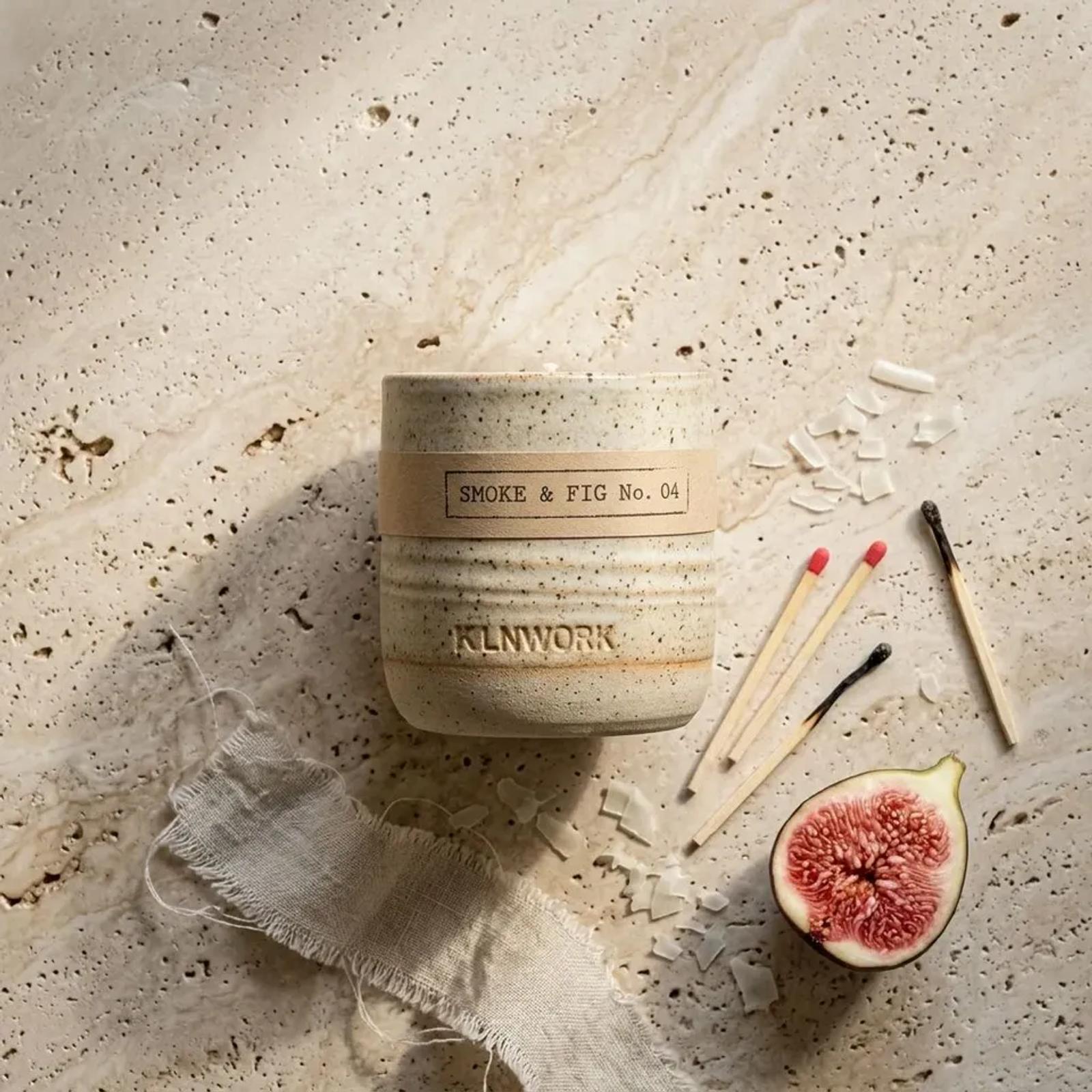 A KLNWORK candle photographed from above on travertine, the paper band reading SMOKE & FIG No. 04 and the maker name pressed into the glaze below it, with spent matches, wax shavings, torn linen and a halved fig around it.