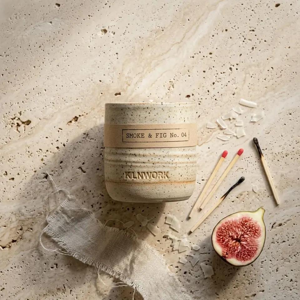 A KLNWORK candle photographed from above on travertine, the paper band reading SMOKE & FIG No. 04 and the maker name pressed into the glaze below it, with spent matches, wax shavings, torn linen and a halved fig around it.