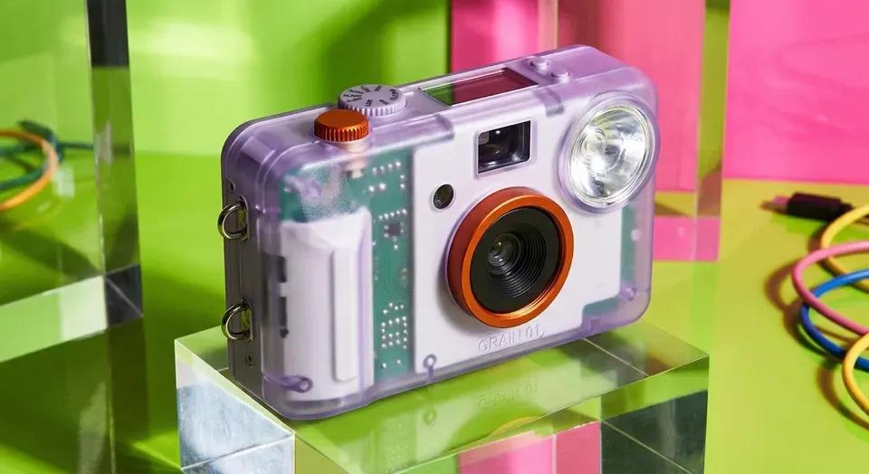 The GRAIN 01 compact camera standing on an acid-green and pink set, translucent lilac body, orange lens ring and a hard flash unit beside it.