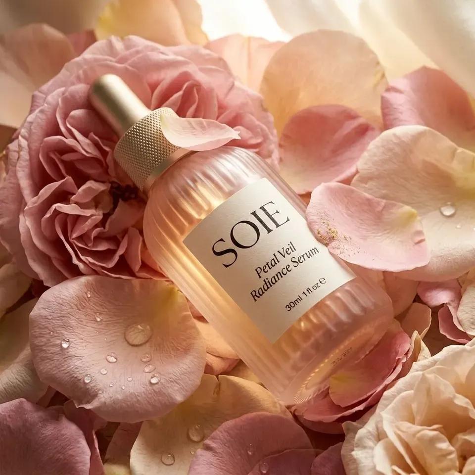 The SOIE bottle lying on its side in a bed of pale pink rose petals beaded with water, the gold cap catching the light.