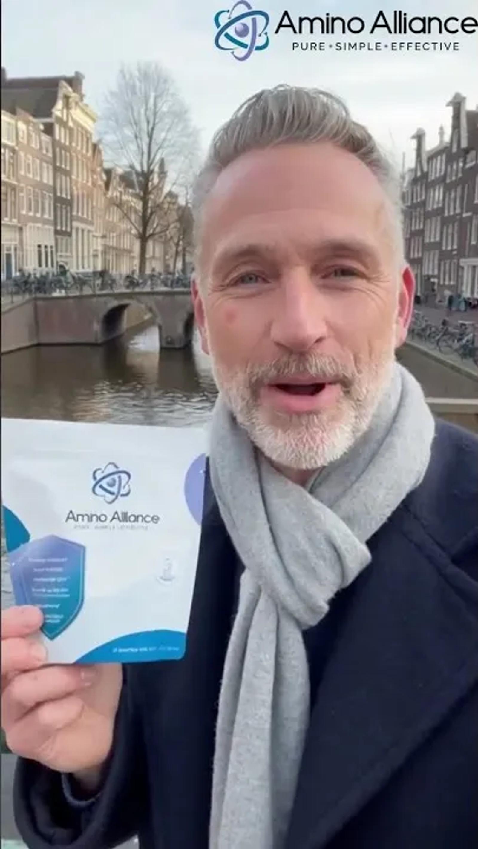 A man in his fifties with grey hair and a grey beard, in a navy overcoat and a pale scarf, holding an Amino Alliance pouch to camera on an Amsterdam canal with a bridge and canal houses behind him.