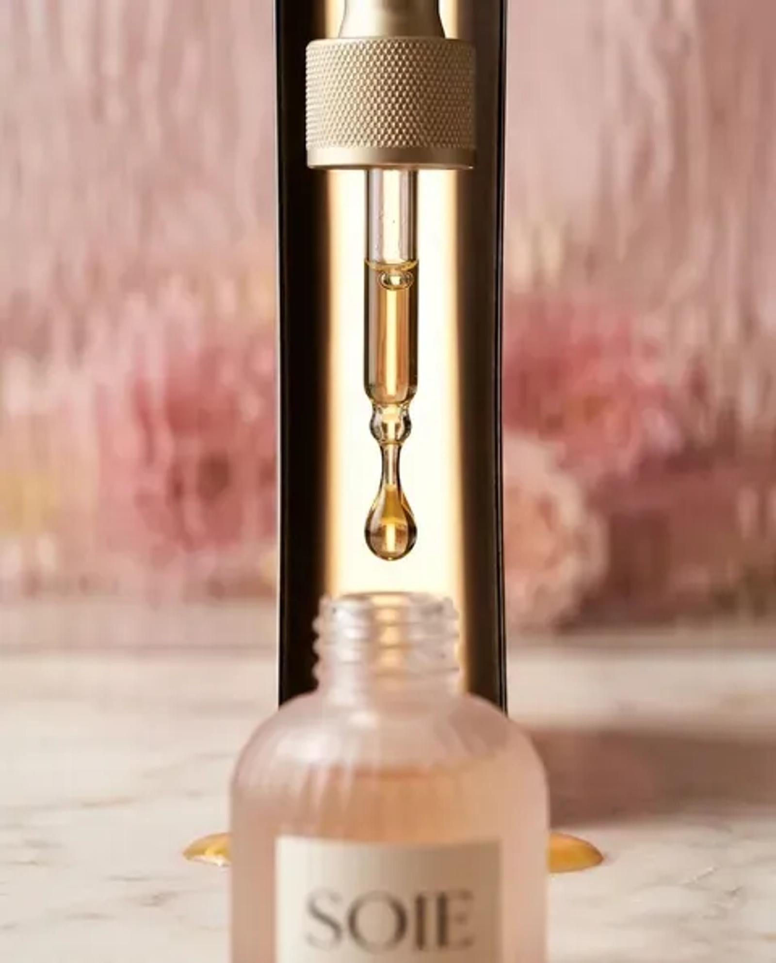 A macro of the SOIE pipette above the open bottle, a single amber drop of serum hanging from the glass tip against a soft pink background.