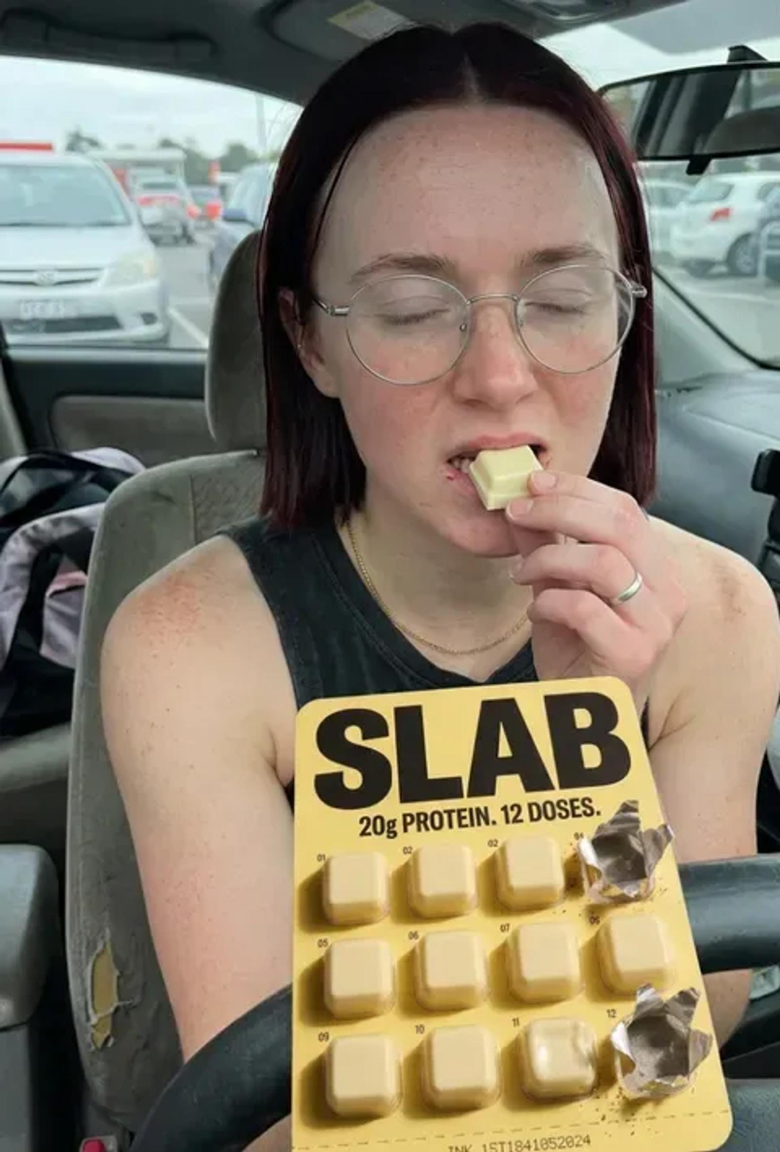 A woman in a parked car biting into a SLAB block, the half-empty tray resting against her chest.