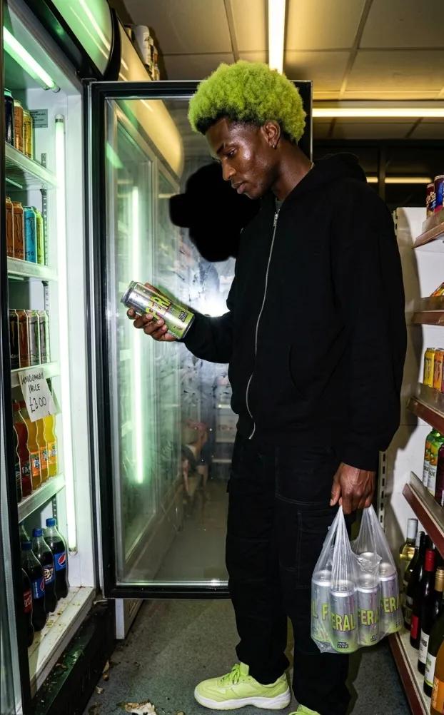 A man with bleached green hair in a black tracksuit reading the side of a FERAL can in front of a lit shop fridge, carrier bags in his other hand.
