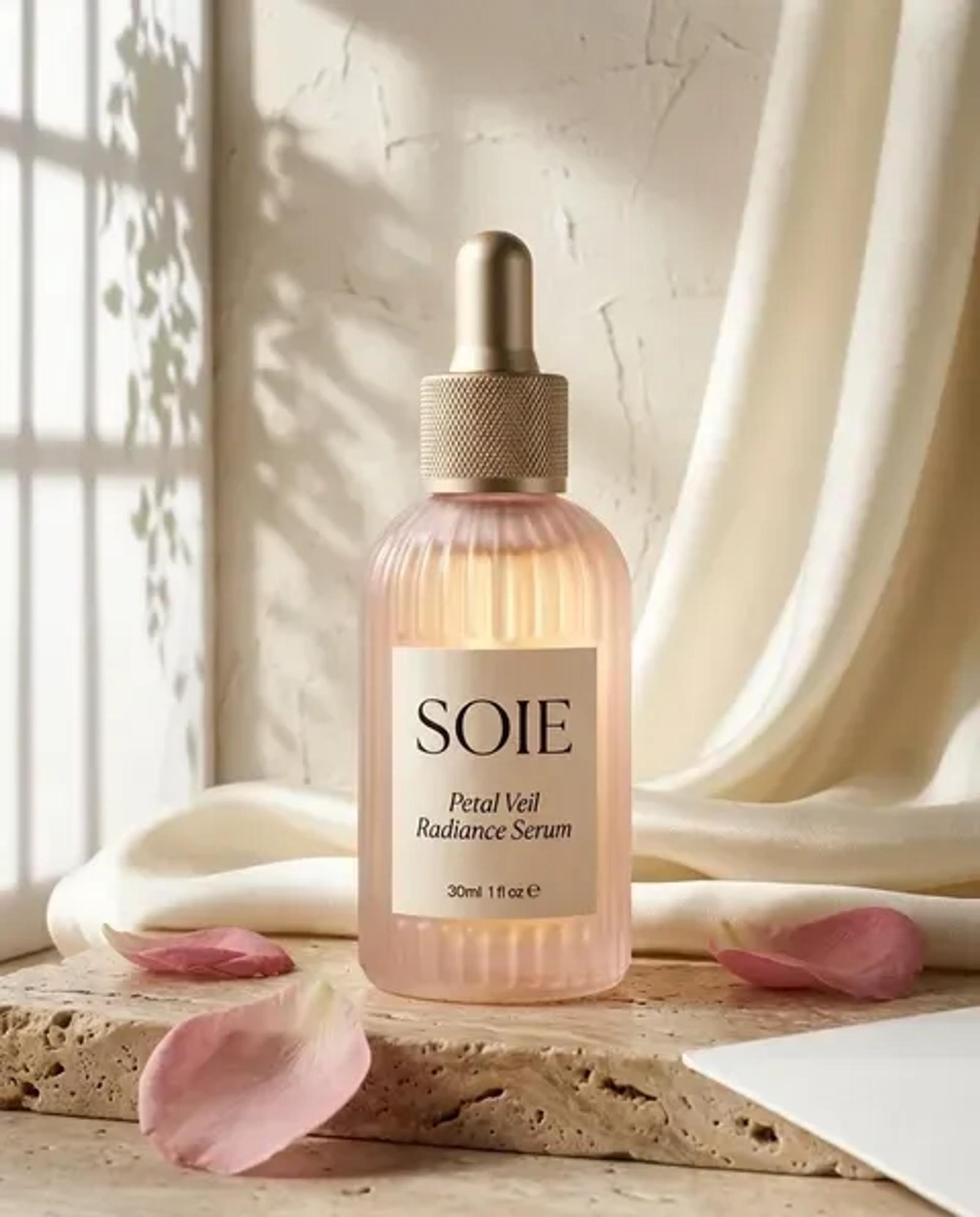 A SOIE Petal Veil Radiance Serum bottle standing on a travertine plinth in front of a linen curtain, rose petals at its base and hard window light raking across the wall behind.