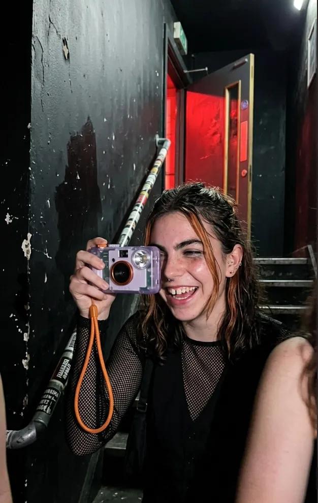 The same woman laughing in a dark club stairwell, raising the compact camera to her eye, red light behind her.