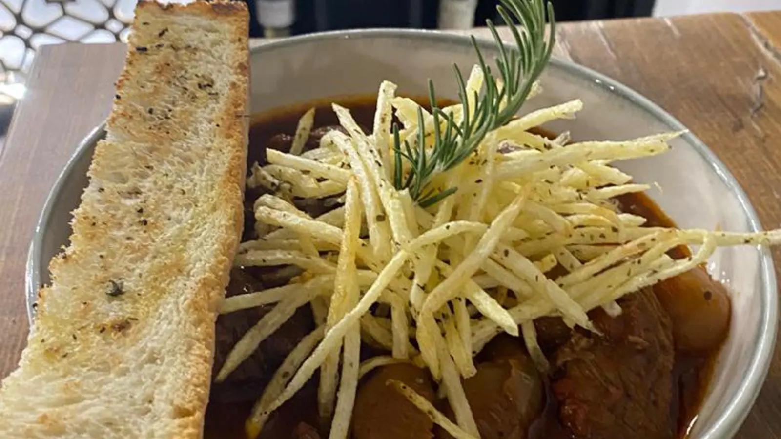 Beef stifado in a dark sauce in a metal bowl under a nest of shoestring fries and a sprig of rosemary, a long slice of grilled bread beside it.