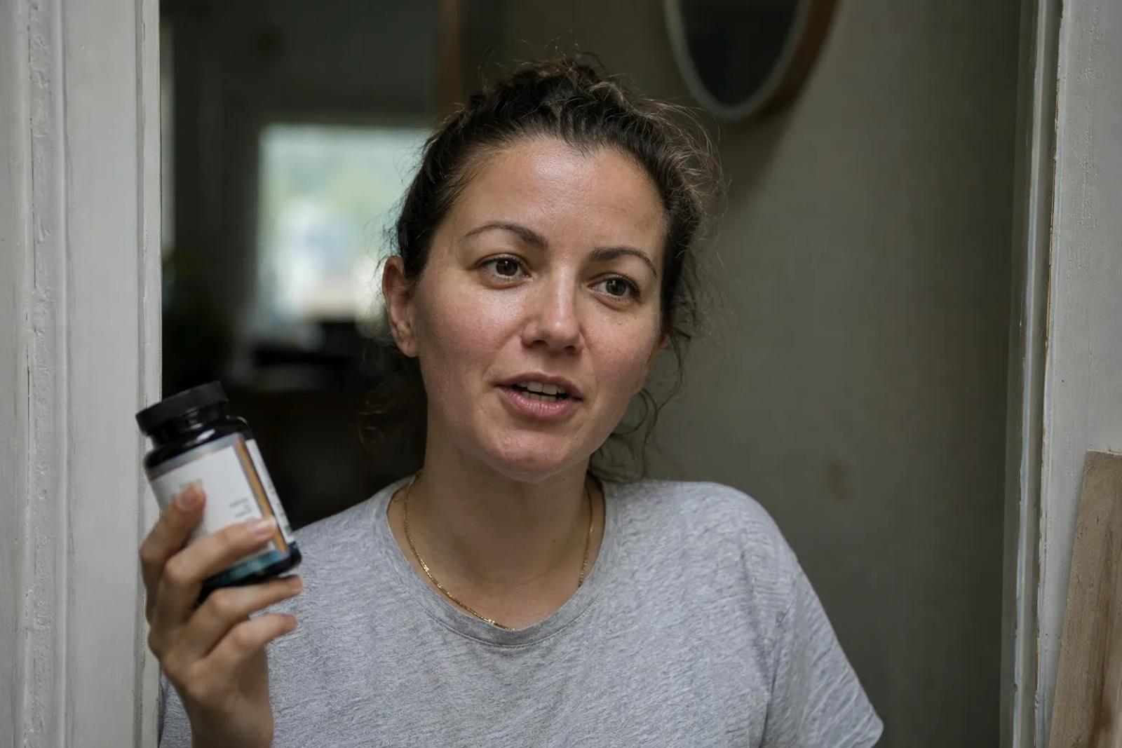 A woman in a grey t-shirt stands in a doorway holding up a dark supplement jar toward the camera, mid-sentence, with an unmade room and a mirror behind her.