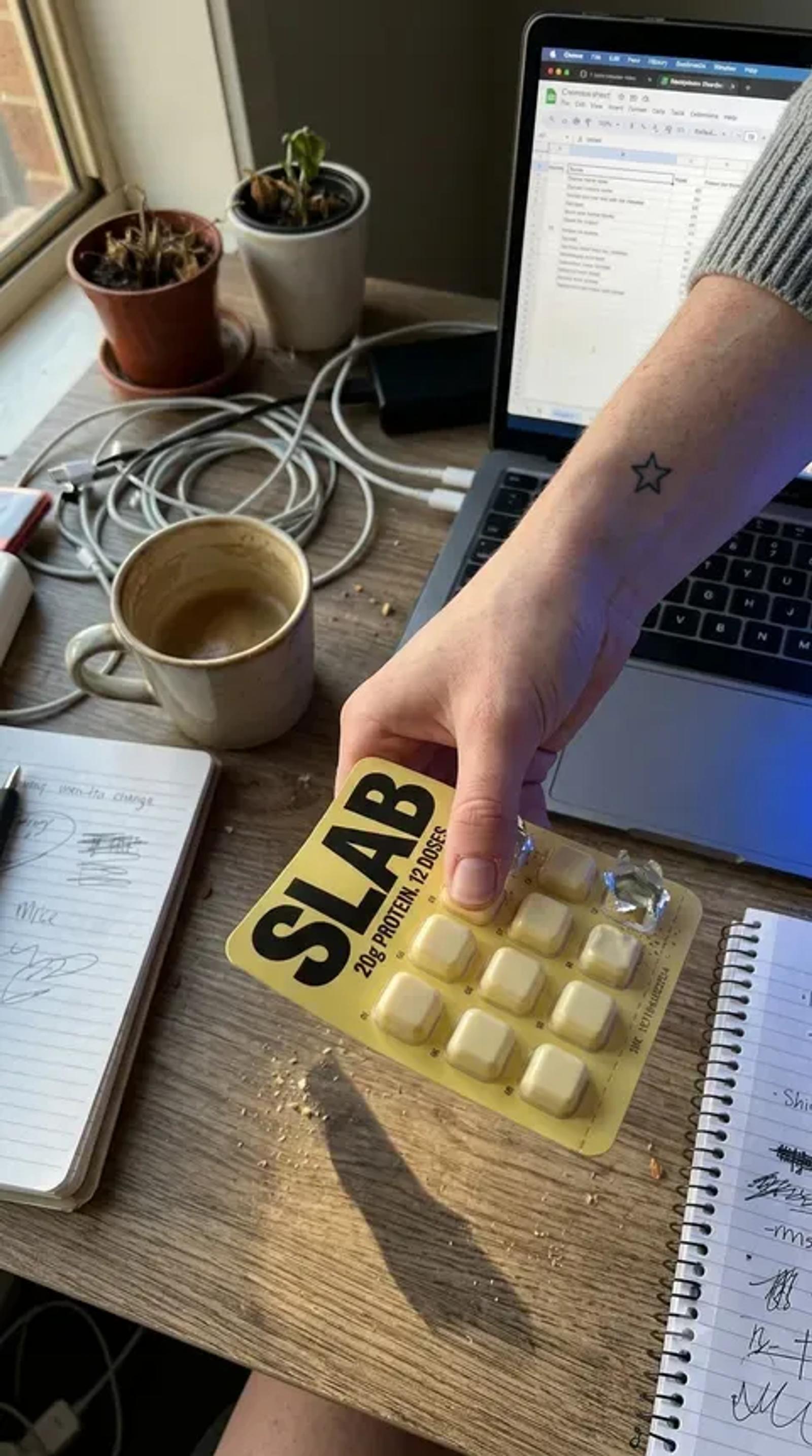 A hand setting a SLAB tray down on a wooden desk beside a laptop, a notebook and a cup of coffee.