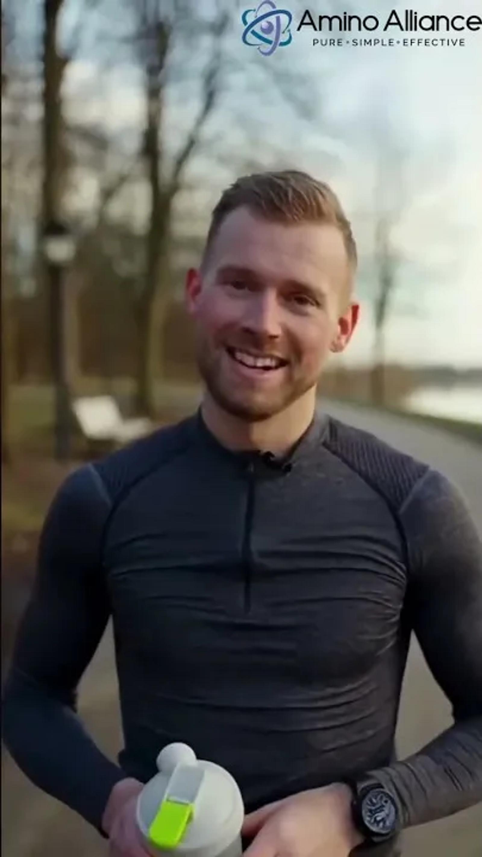 A man in a charcoal quarter-zip running top holding an Amino Alliance shaker, on a park path beside water under bare winter trees.