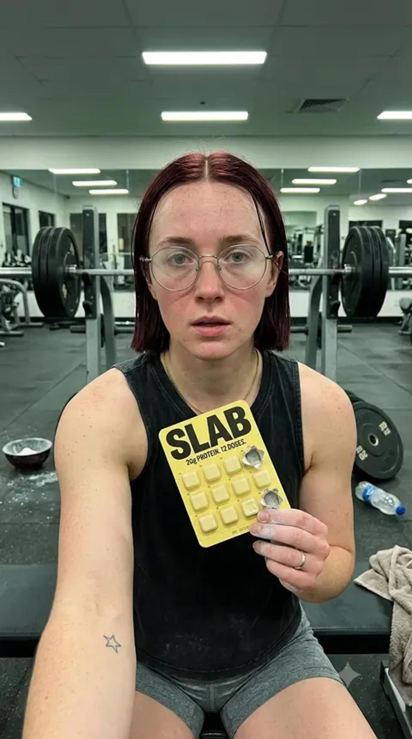 A woman in a gym vest holding a SLAB tray flat to camera, weights racked behind her under strip lighting.