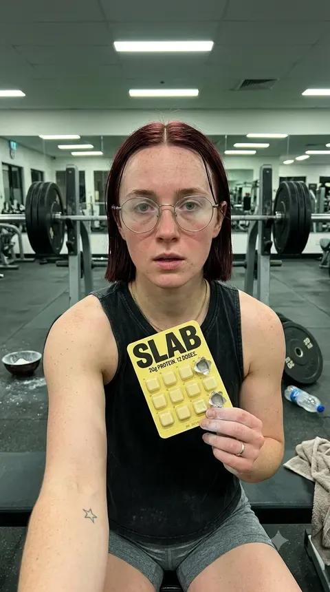 A woman in a gym vest holding a SLAB tray flat to camera, weights racked behind her under strip lighting.