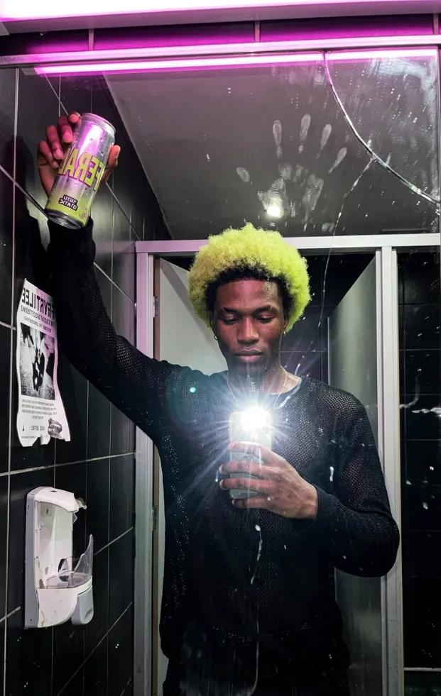 The same man holding a FERAL can up in a bathroom mirror selfie, flash on, pink strip light above the cracked mirror.