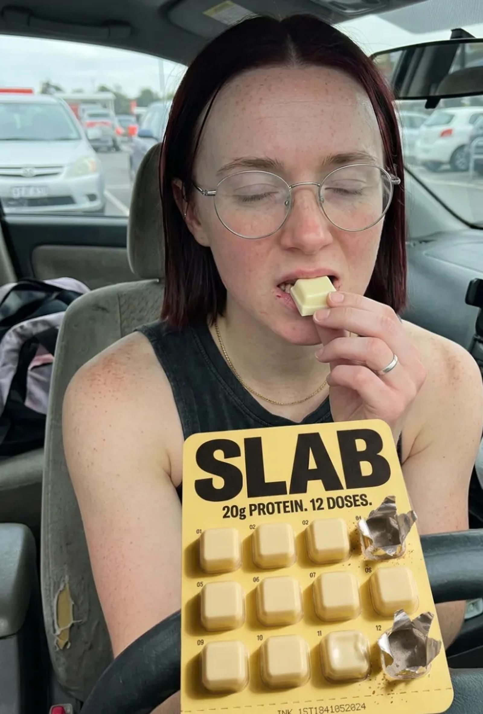 A woman in a car park eating a SLAB block, holding the yellow tray up to camera with the pack claim facing forward.