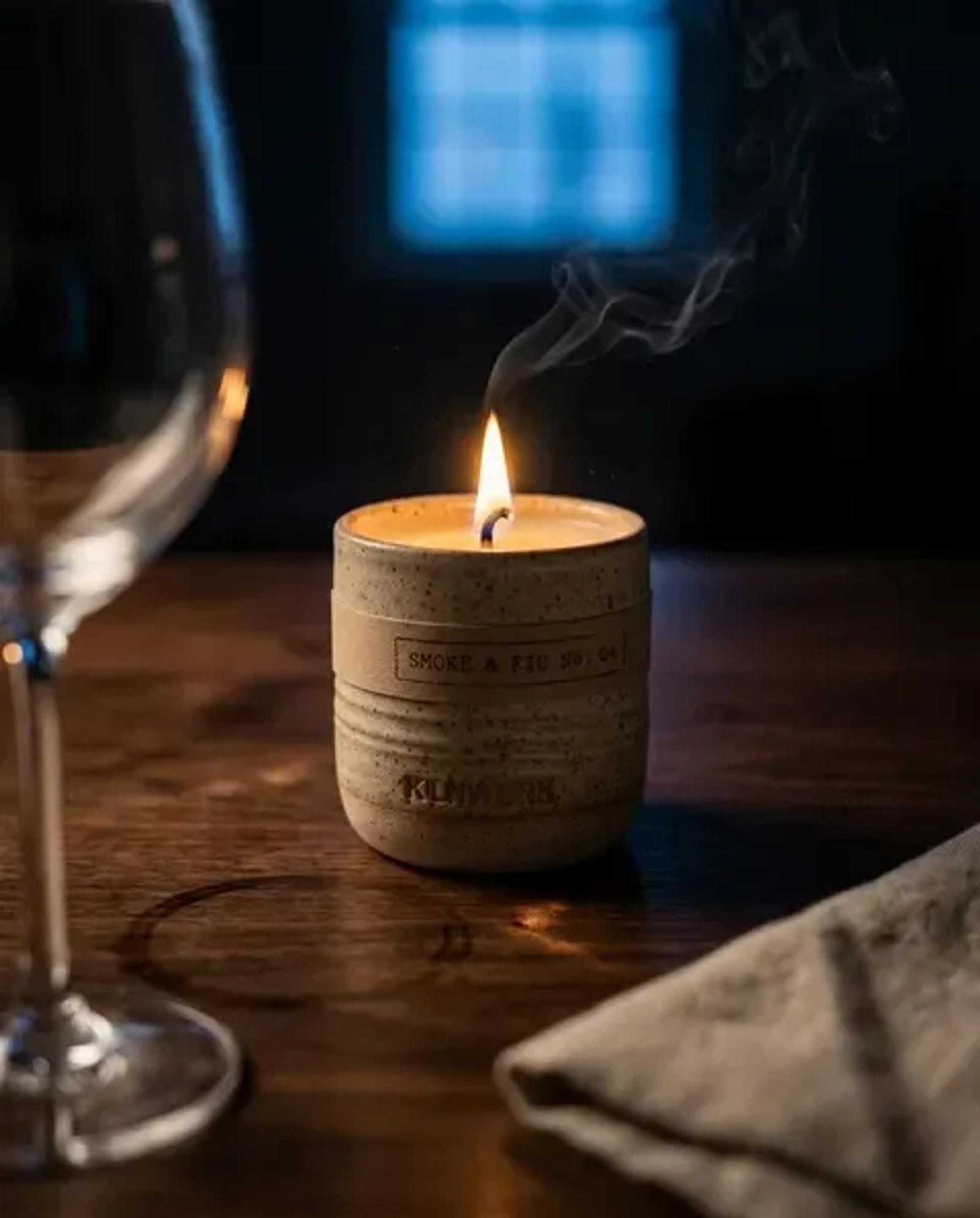 The same candle lit in a dark room, a thin smoke trail rising from the wick, an empty wine glass and a linen napkin catching the flame light beside it.