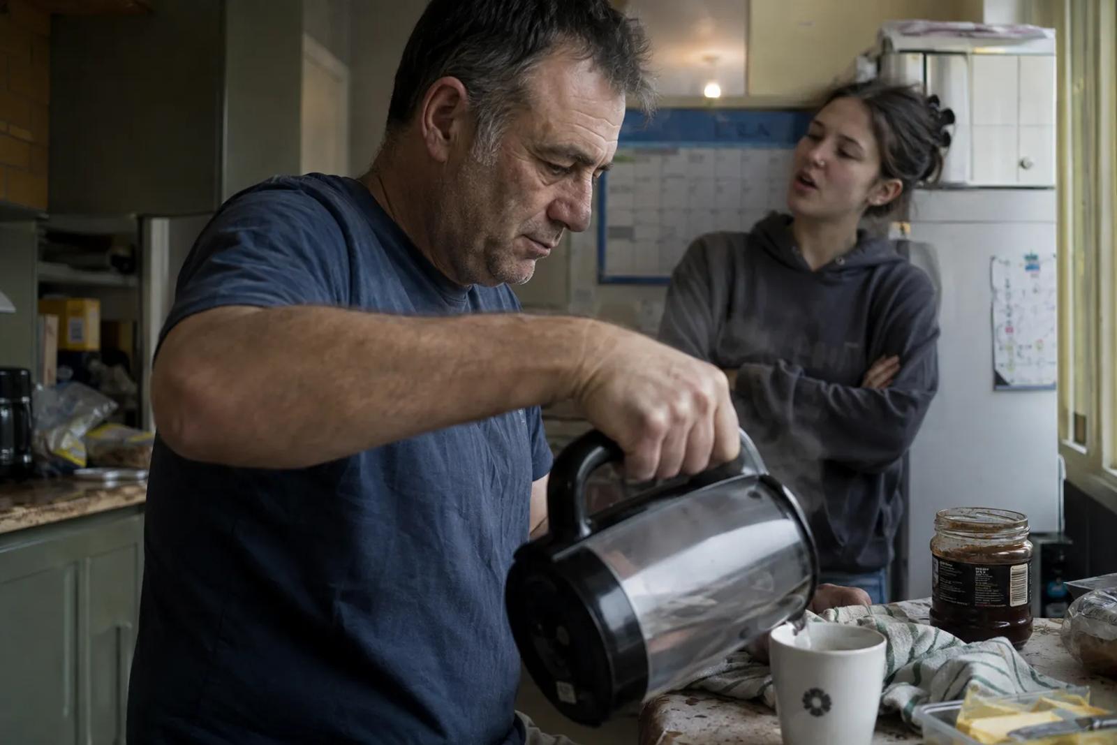 A man in a navy t-shirt pours from a glass coffee jug into a mug in a cluttered domestic kitchen while a young woman in a grey hoodie stands behind him with her arms folded, mid-sentence, looking at him.