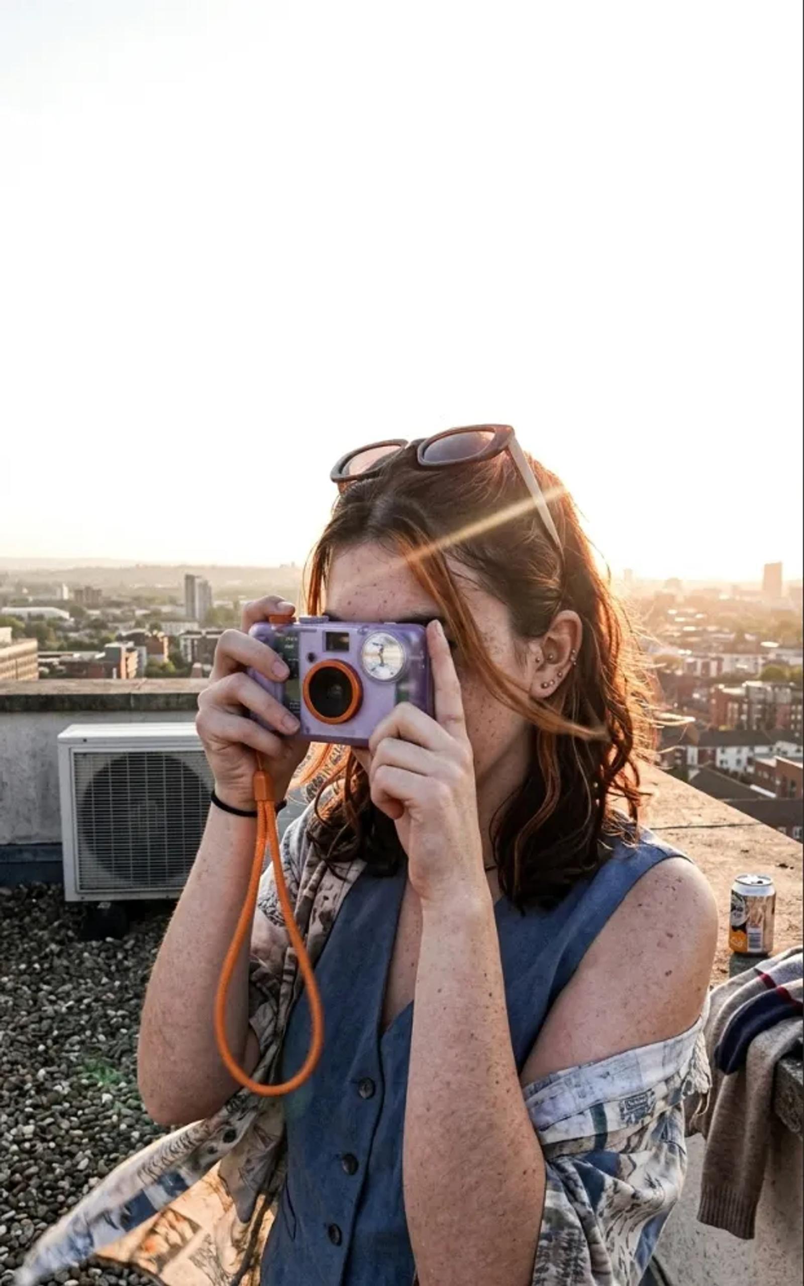 The same woman on a rooftop at sunset, camera to her eye, city and air conditioning units behind her.