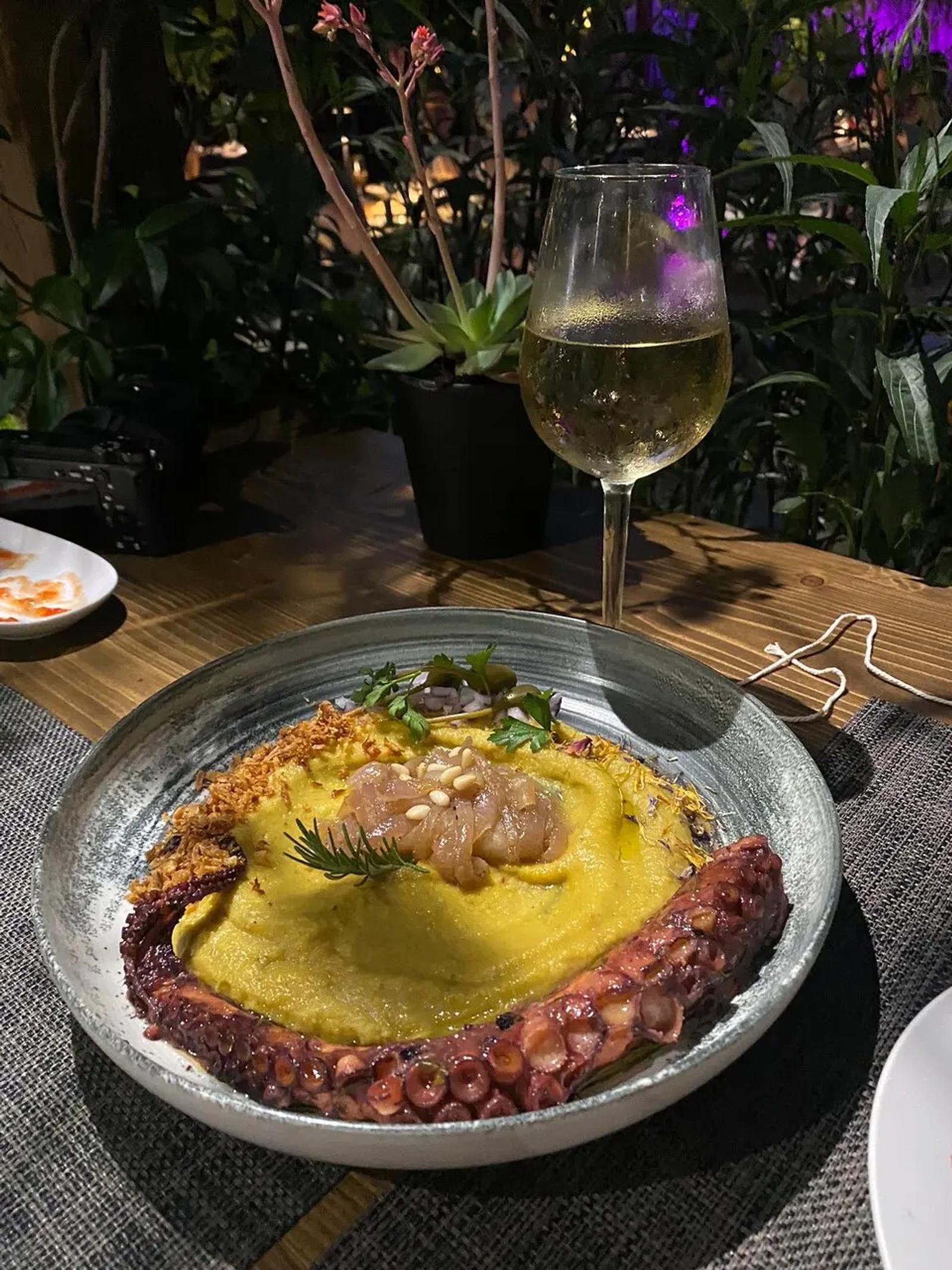 Grilled octopus curled over yellow fava purée in a stone bowl, topped with pickled onion and dill, shot at the table at night with a glass of white wine behind it.