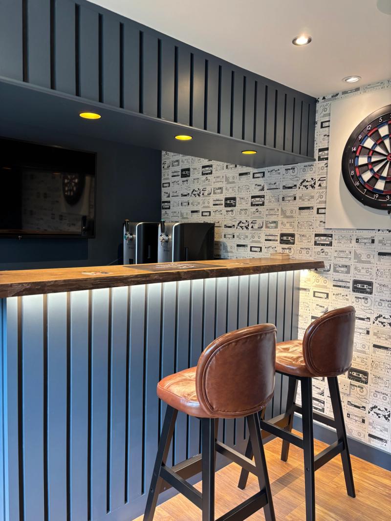The finished home bar in Ewell: navy panelled counter with a walnut top, twin beer taps, a dartboard and cassette-print feature wall.