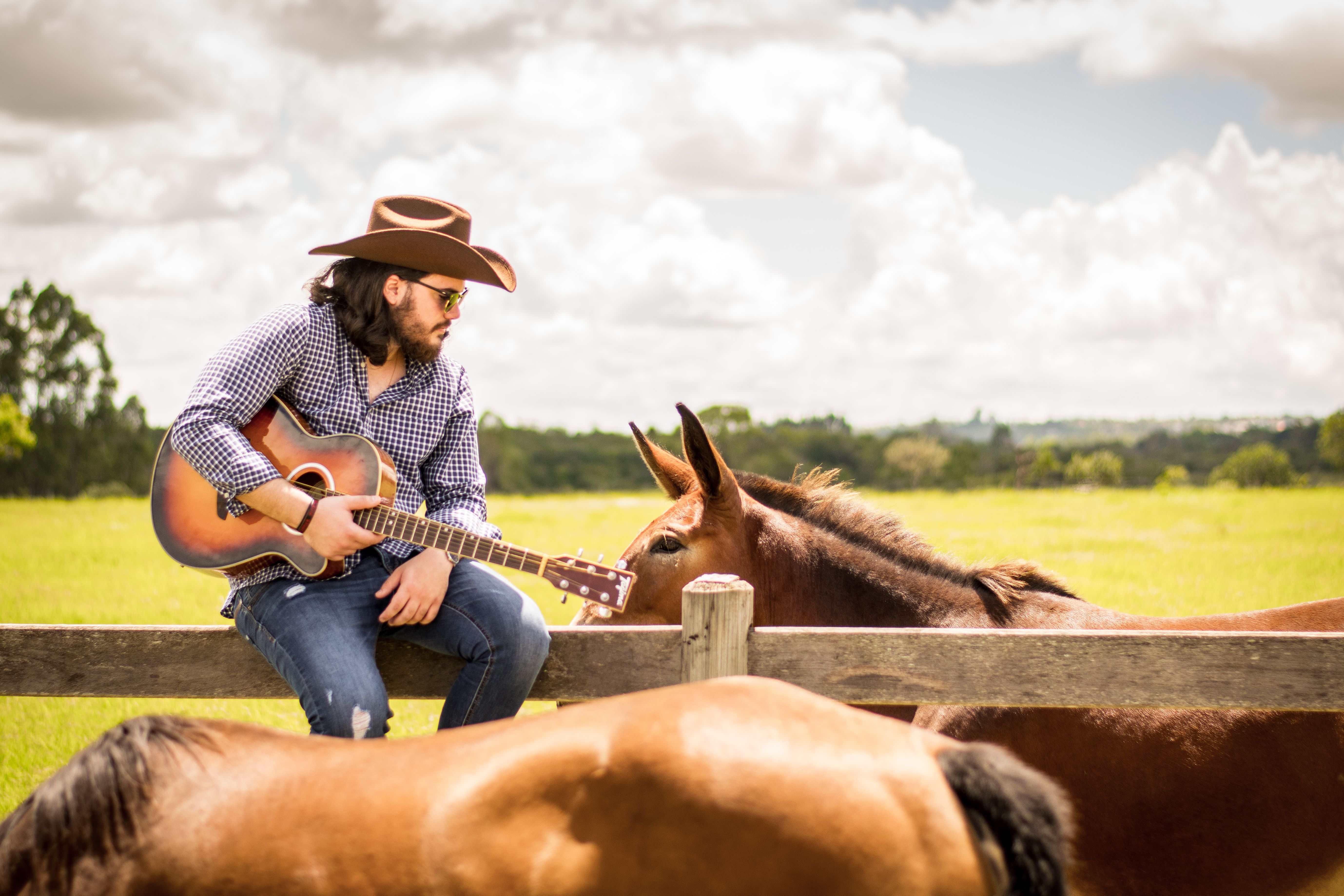 Matheus Granjeiro sentado na cerca com o violão ao lado de um cavalo
