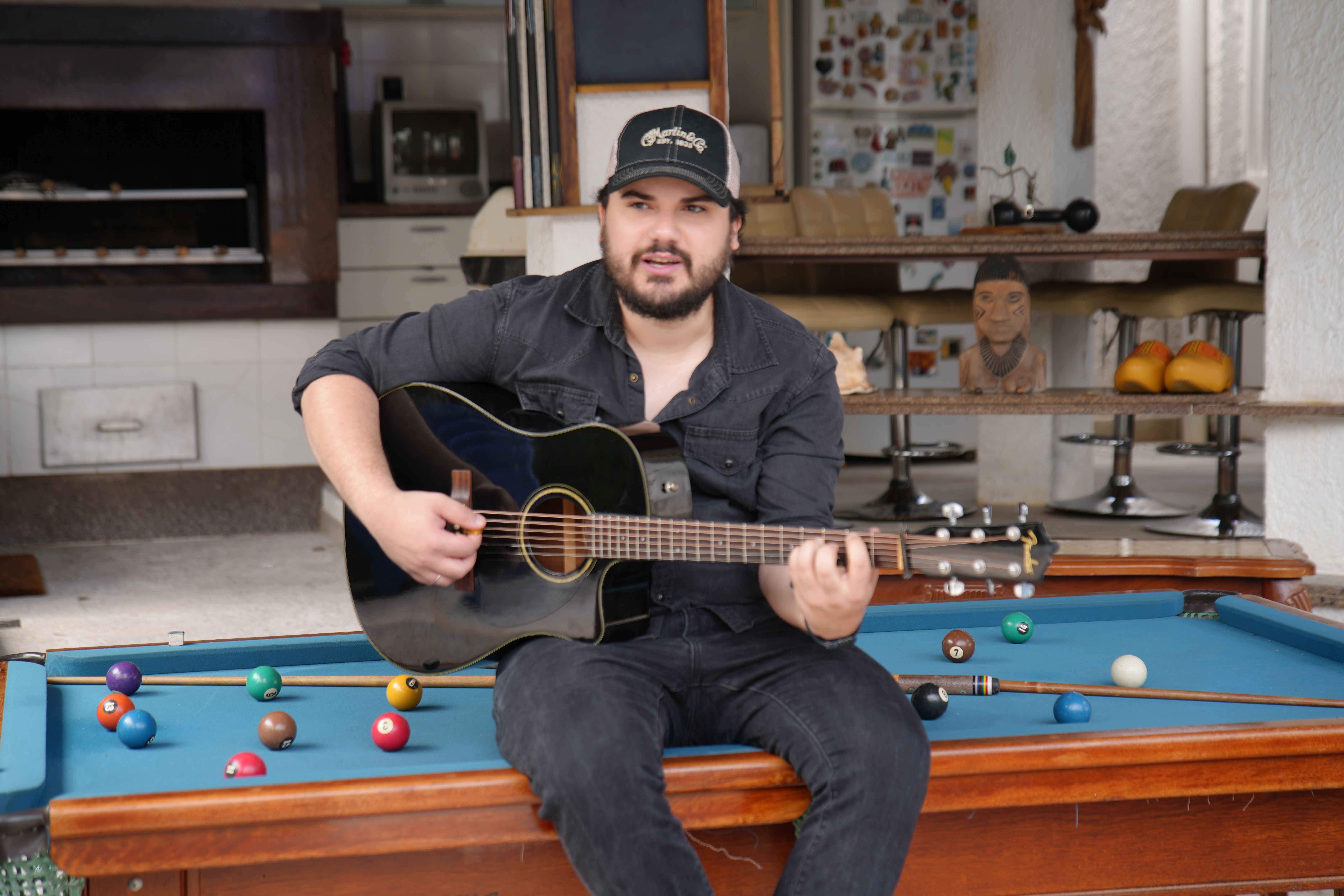 Matheus Granjeiro sentado na mesa de sinuca tocando violão