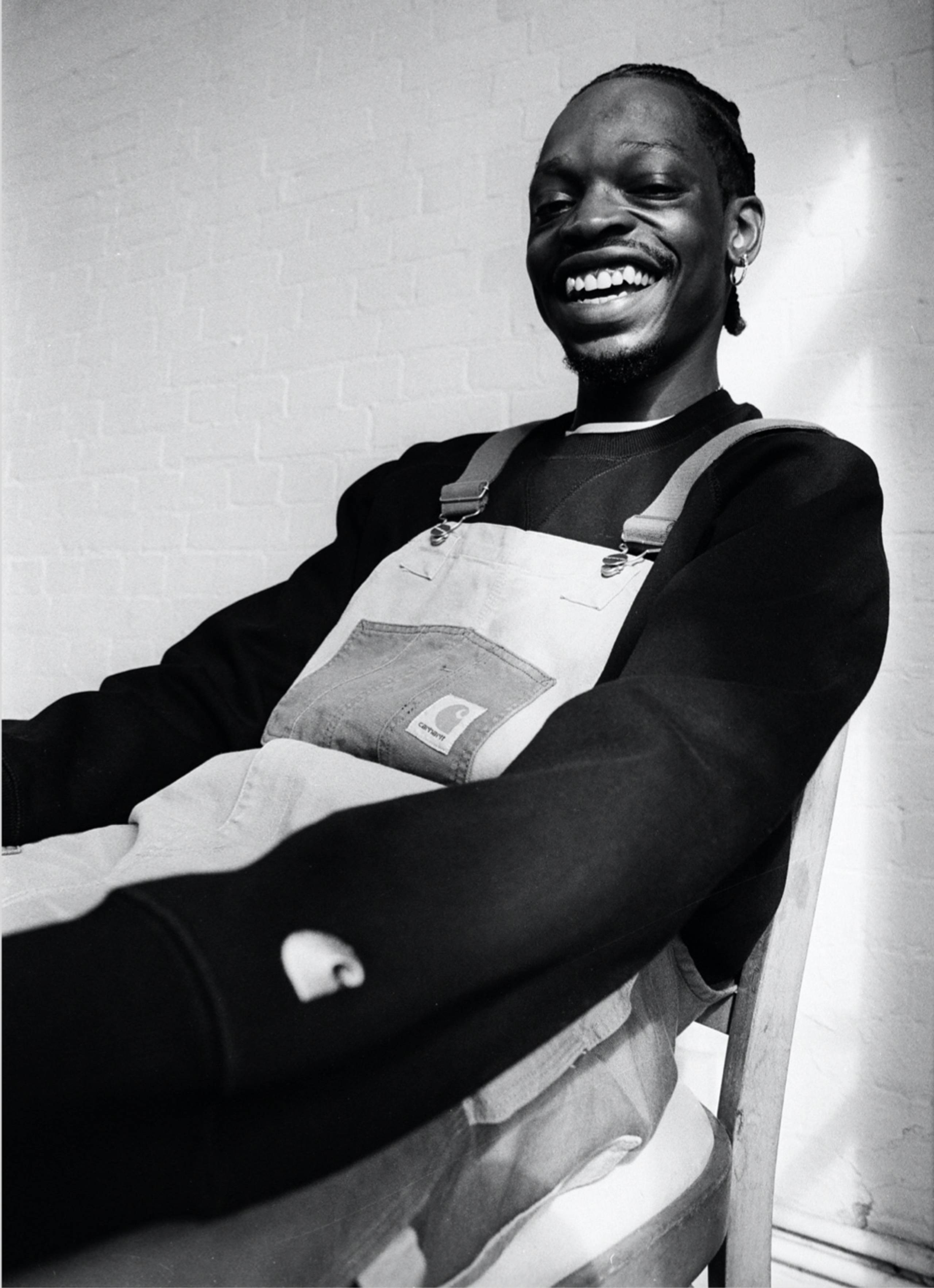 Black and white photo of seated Model wearing Carhartt WIP Double Knee Bib Overall in Two Tone Dusty Hamilton Brown