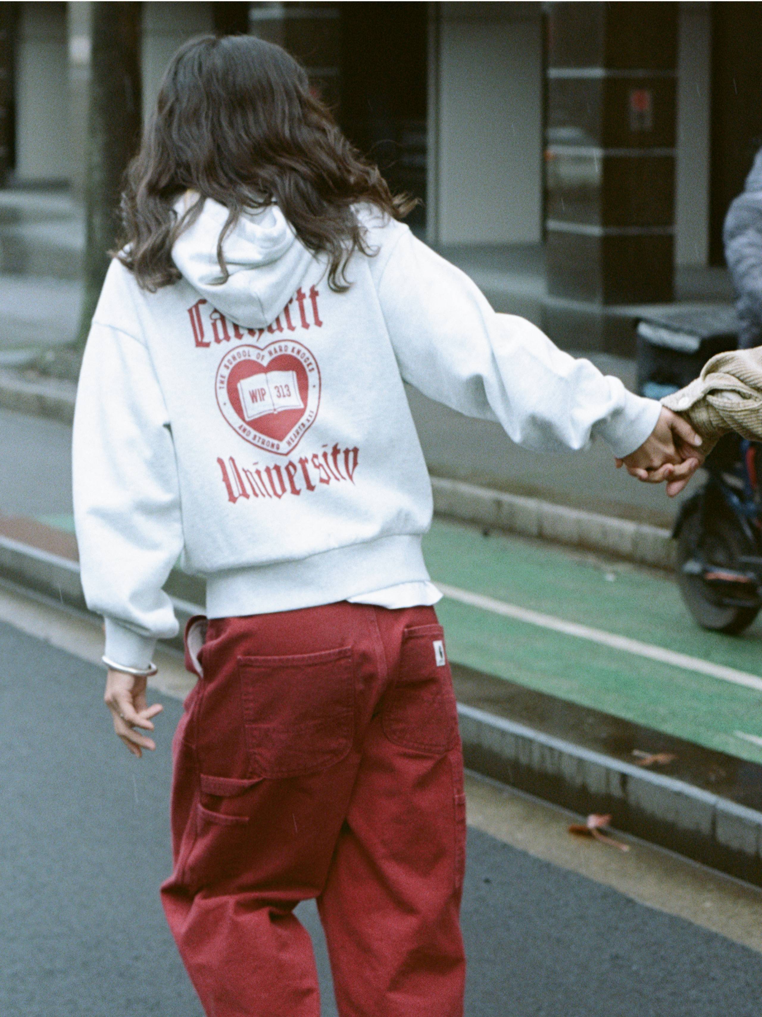 The back of a person wearing a "Carhartt University" hooded sweatshirt and red double knee pants