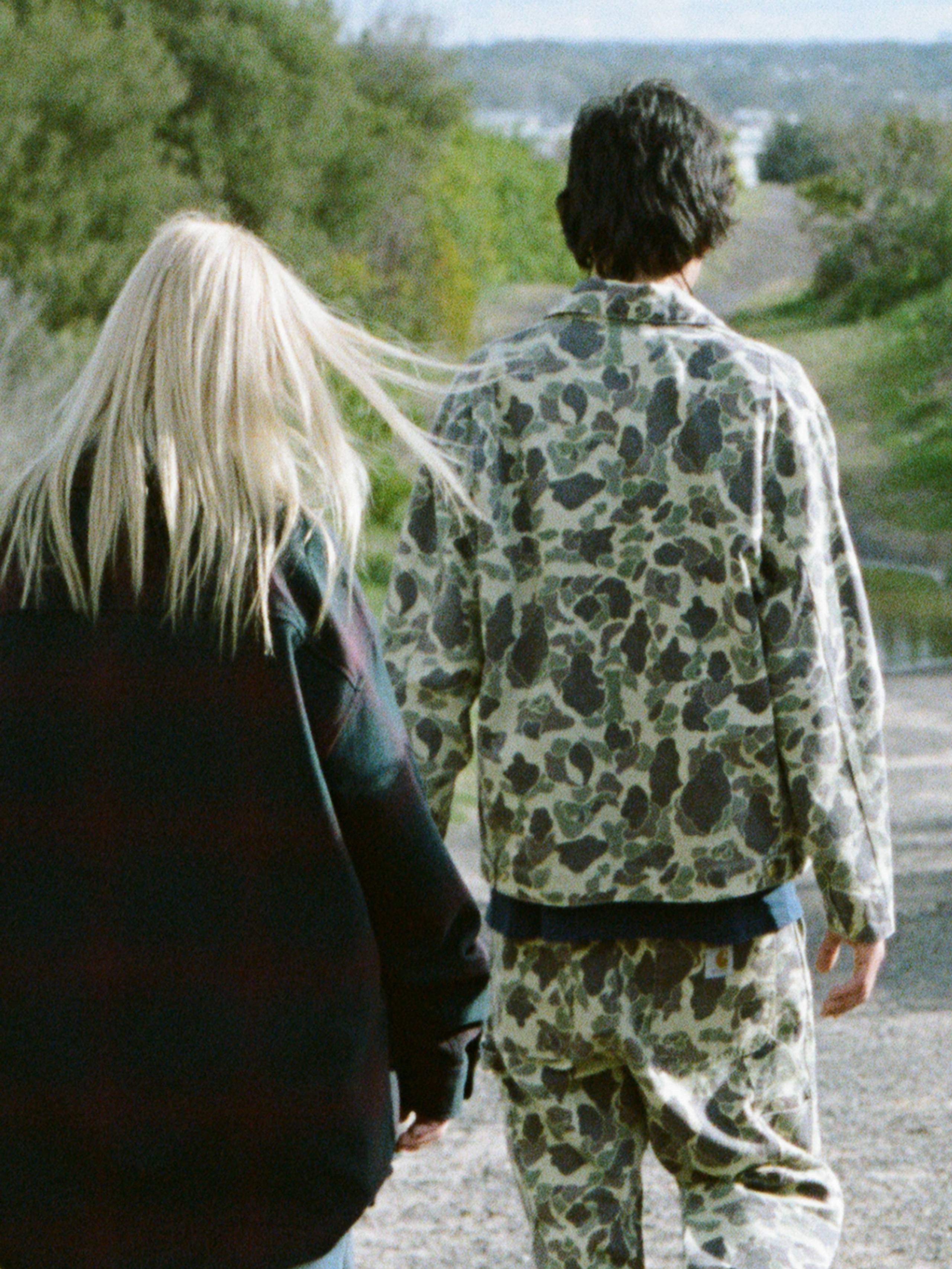 The back view of three people walking down a path, person on the left wearing a black jacket. The person in the middle is wearing camo jacket and pants, the person on the right wearing a denim active jacket.
