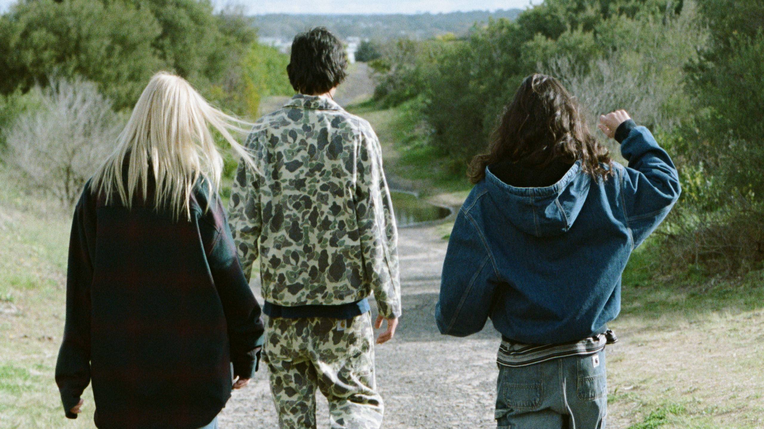 The back view of three people walking down a path, person on the left wearing a black jacket. The person in the middle is wearing camo jacket and pants, the person on the right wearing a denim active jacket.
