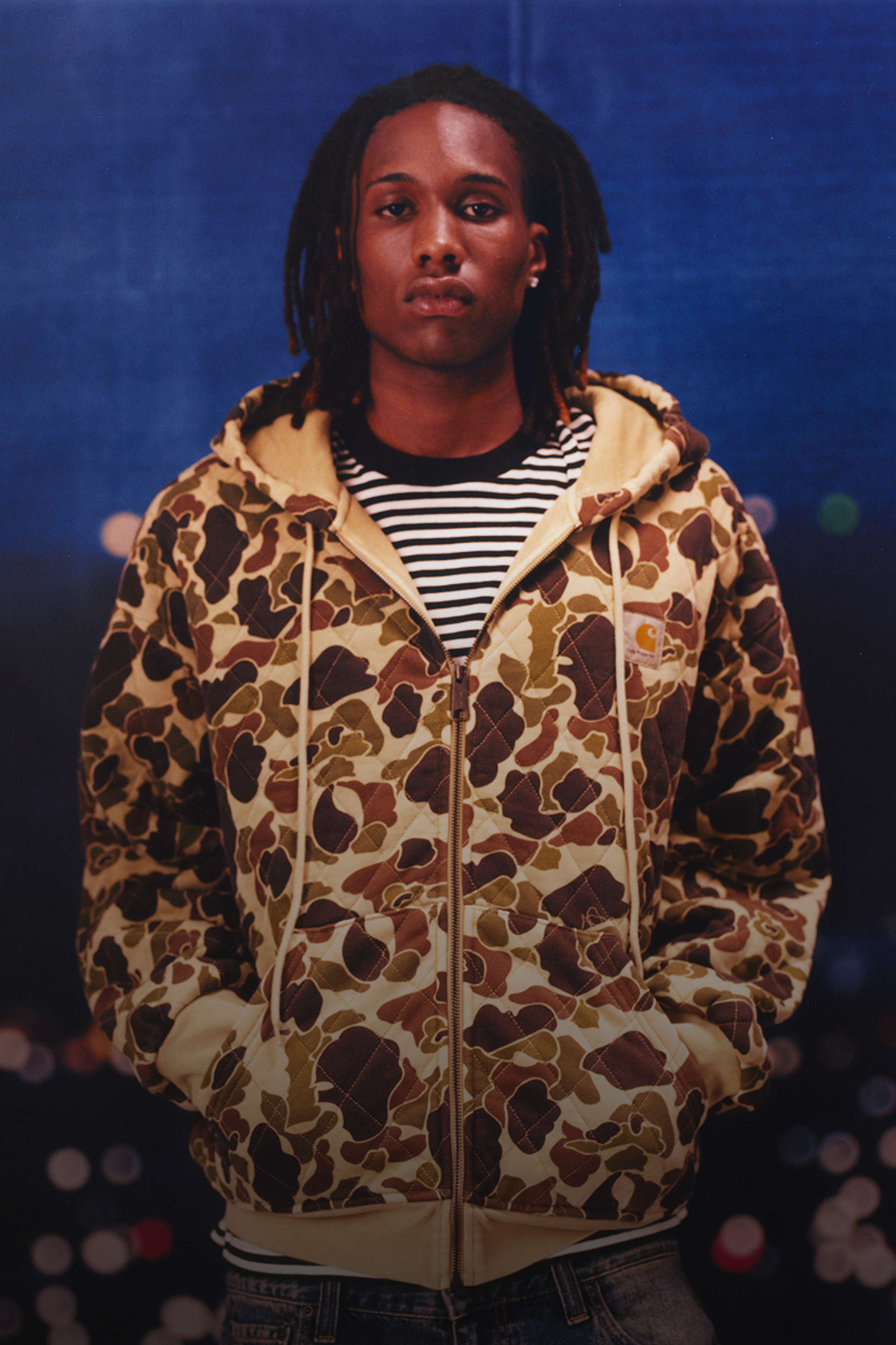 Model wearing a camo hooded sweatshirt, striped t-shirt and light washed denim in the Carhartt WIP F/W25 Men's Lookbook