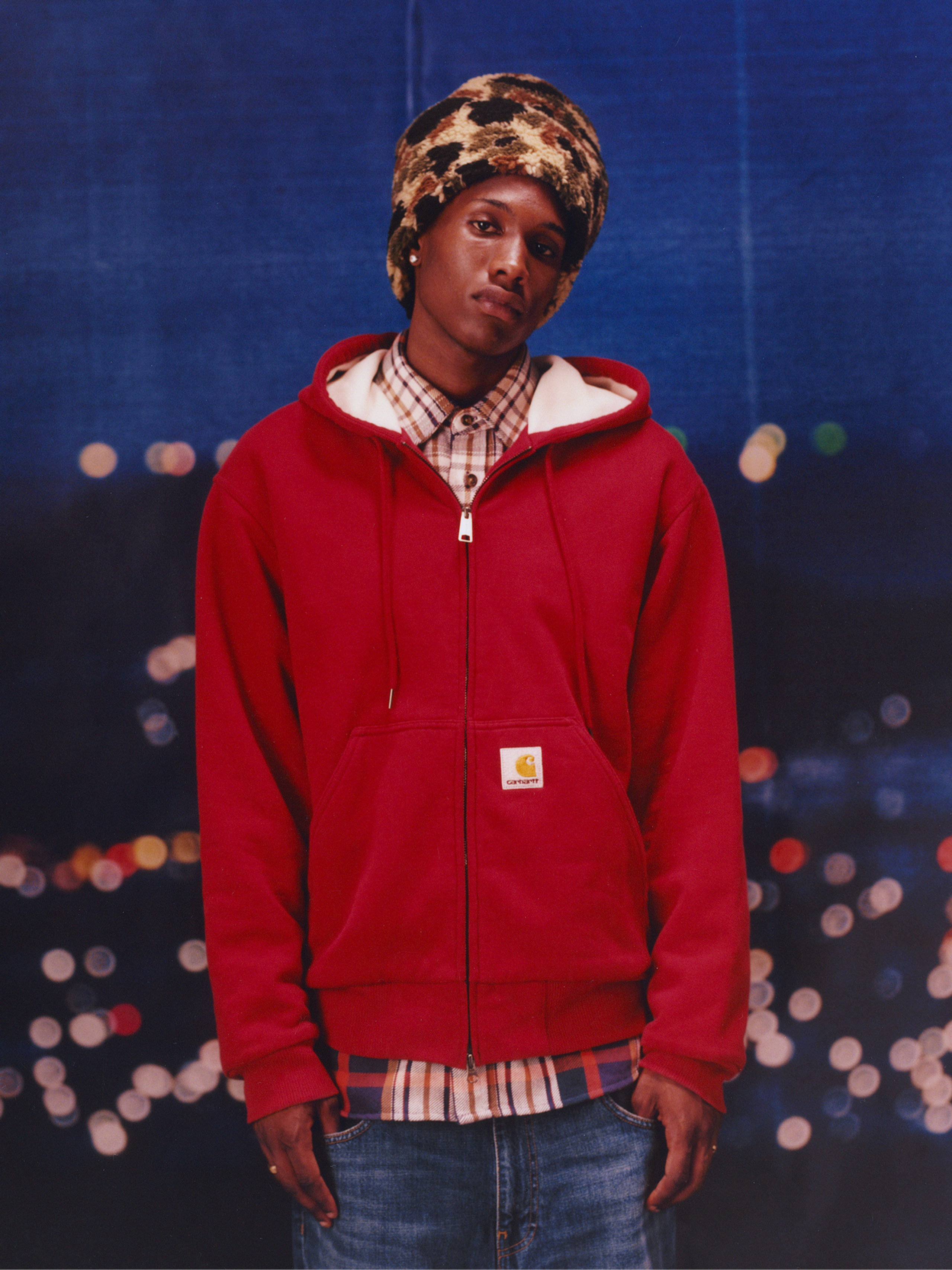Model wearing red sweatshirt and camo beanie in the Carhartt WIP F/W25 Men's Lookbook