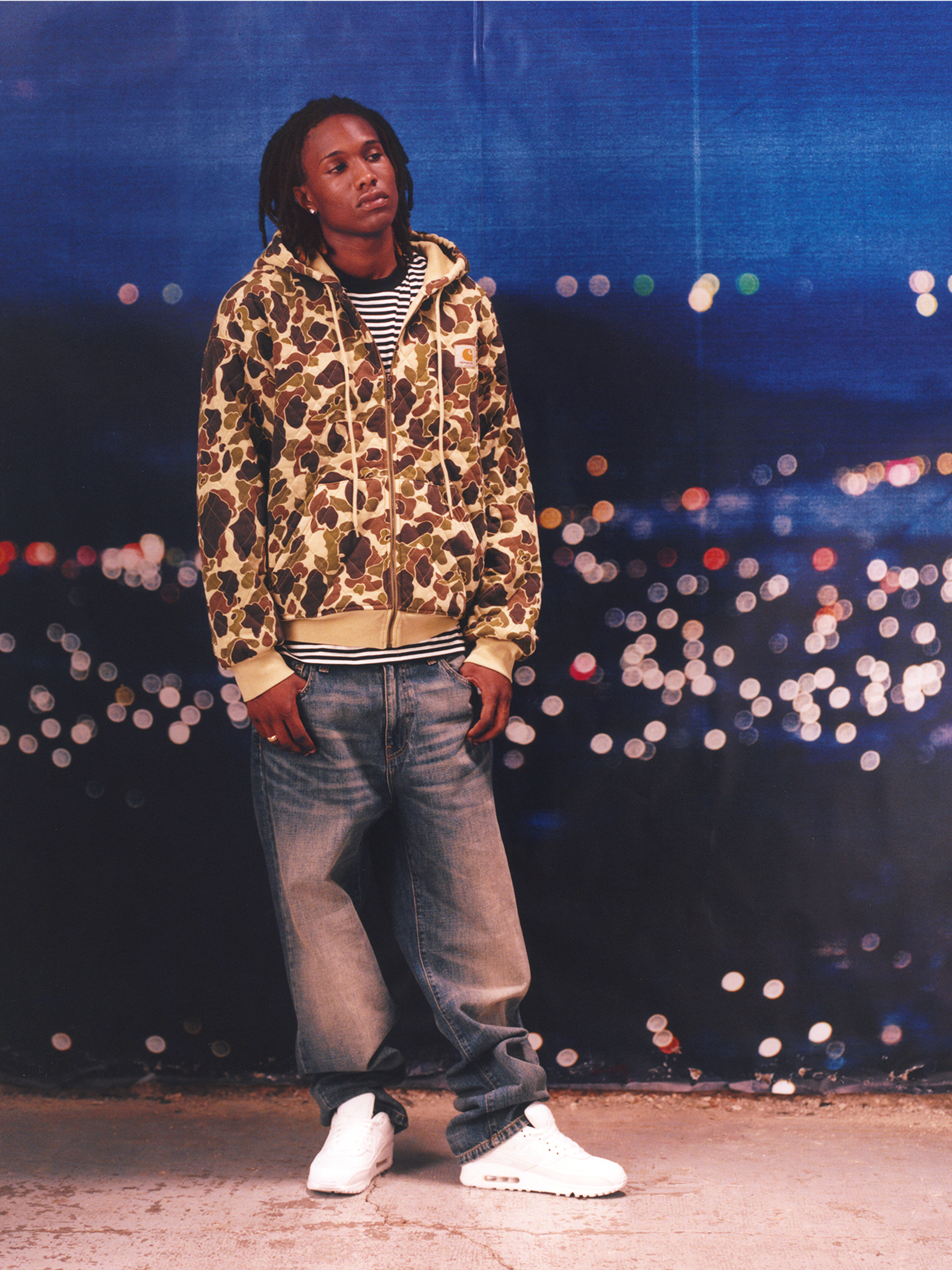 Model wearing a camo hooded sweatshirt and light washed denim in the Carhartt WIP F/W25 Men's Lookbook